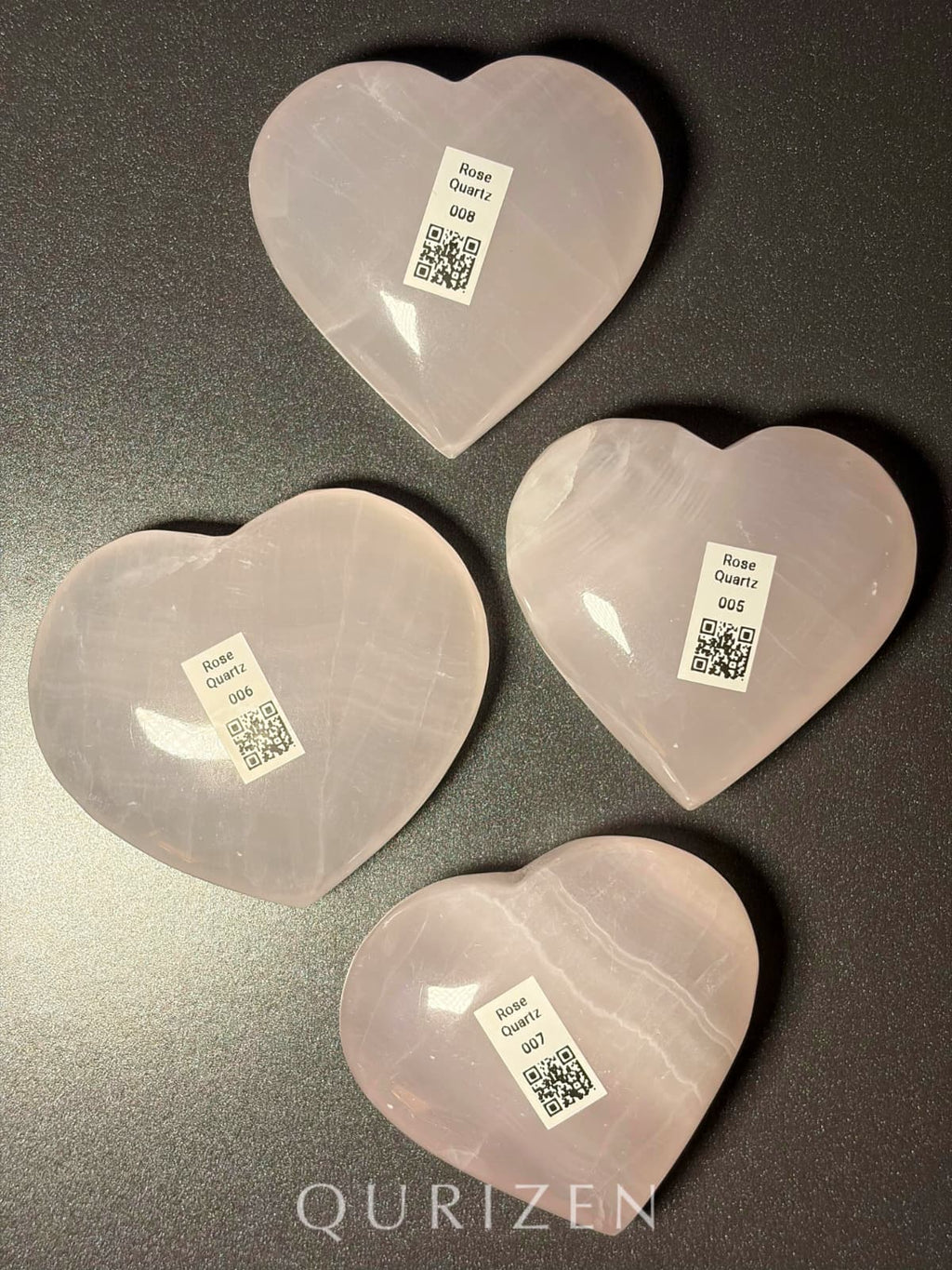 Four heart-shaped rose quartz crystals with QR code labels on a dark surface, branded 'QURIZEN'.