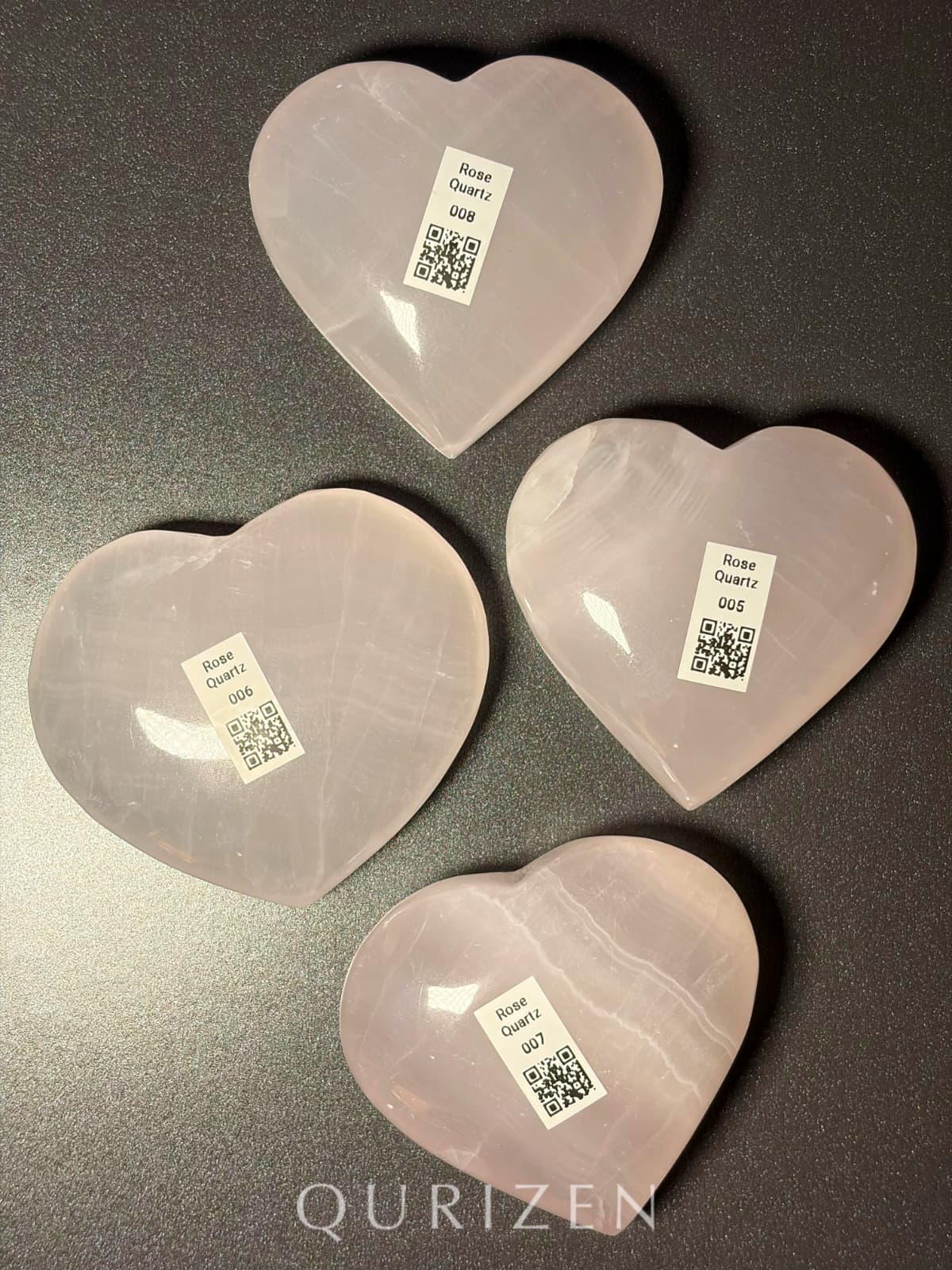 Four heart-shaped rose quartz crystals with QR code labels on a dark surface, branded 'QURIZEN'.