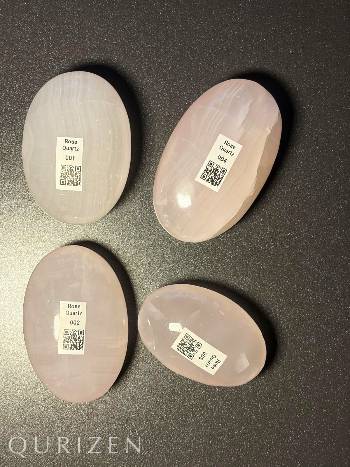 Four rose quartz stones with labels on a dark surface, branded 'Qurizen'.