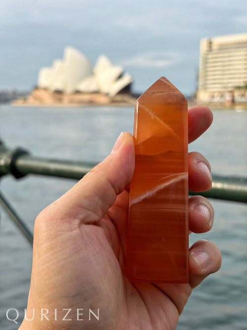 Hand holding a polished orange crystal with a scenic background of a body of water and buildings.