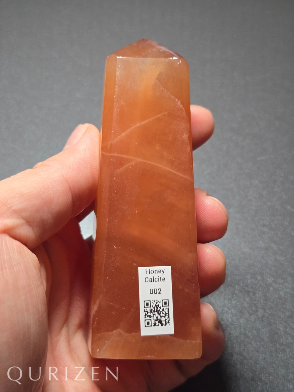 Honey calcite crystal held in a hand with a QR code label, against a gray background.