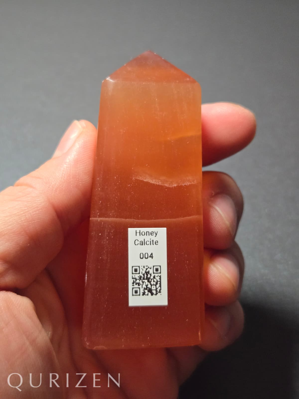 Honey calcite crystal held in a hand with a QR code label, against a gray background.