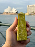 Hand holding a green crystal with a blurred background of a famous landmark and water.