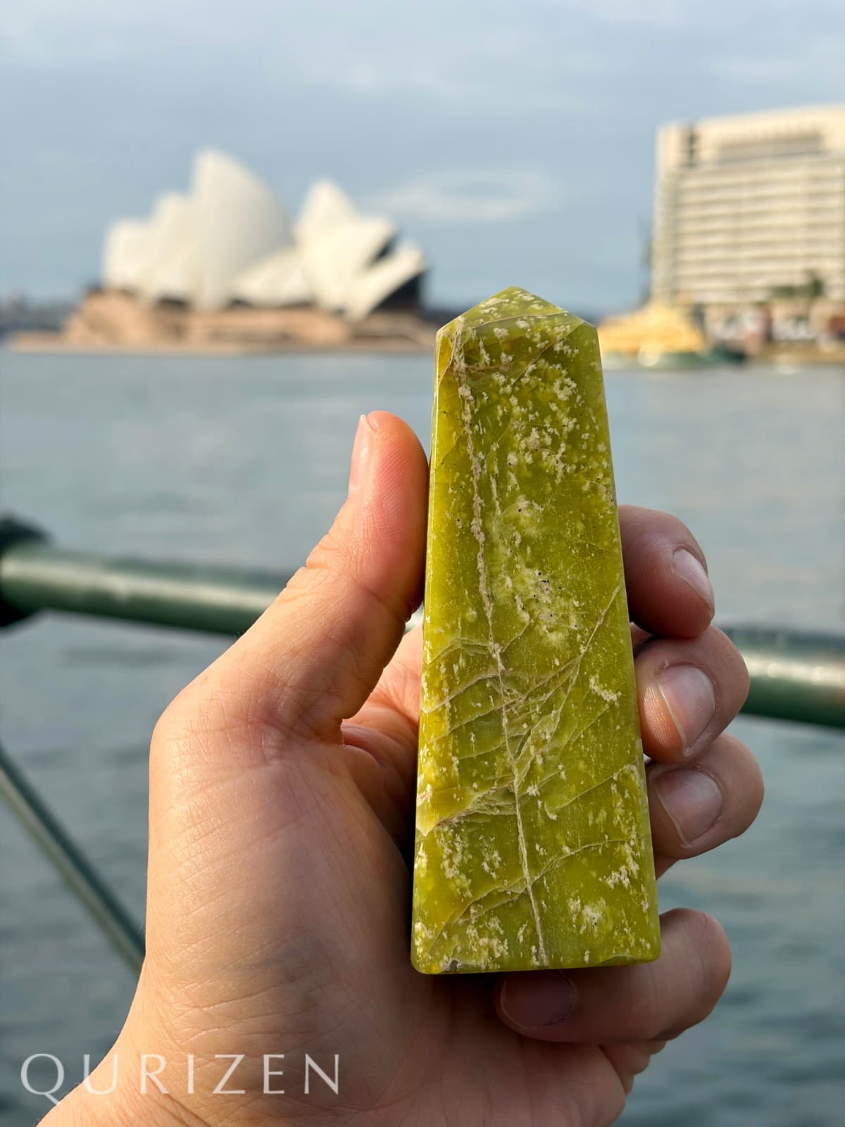 Hand holding a green crystal with a blurred background of a famous landmark and water.